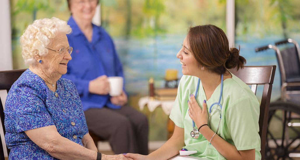 Latin, female doctor or nurse conducts family consultation with elderly patient and her daughter (background) in a nursing home or clinic setting.  Woman is over 100 years old!